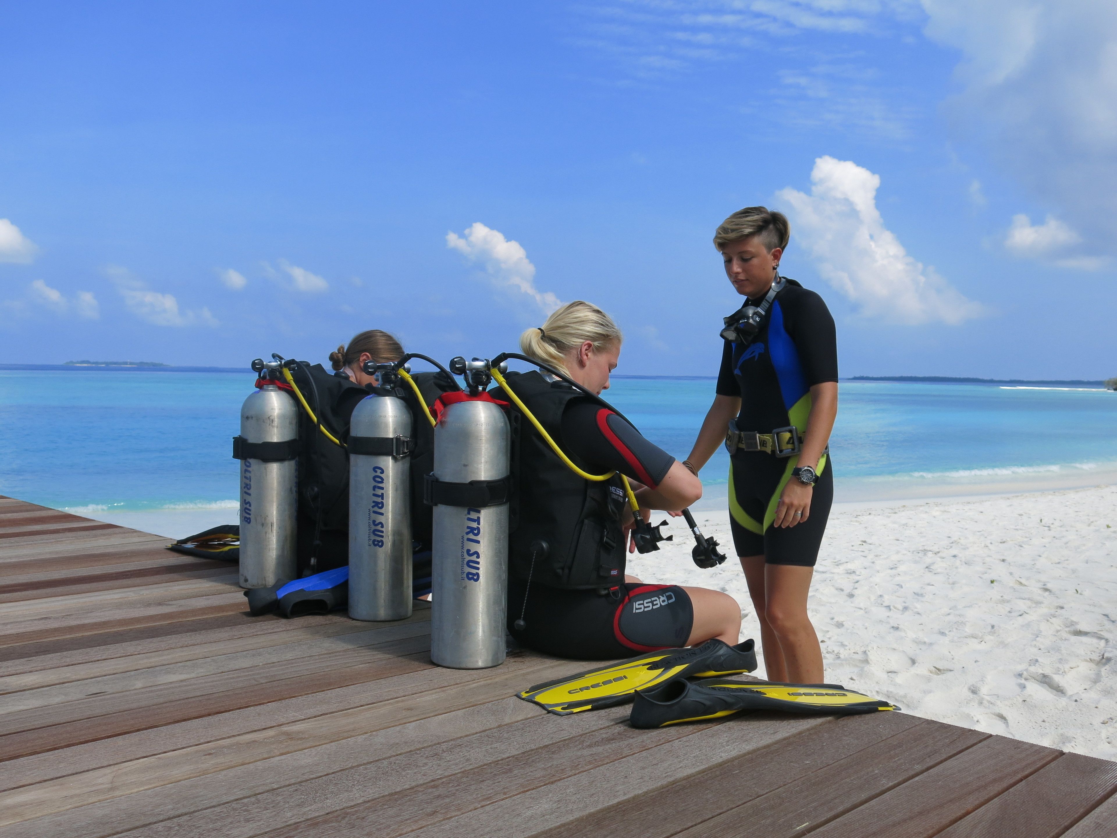 A wooden deck with three scuba tanks labeled "COLTRISUB" and a diver's gear on the sand.