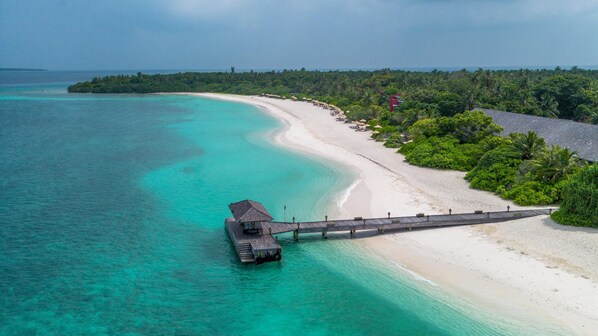 Una vista aerea di una spiaggia con un molo di legno che conduce a un piccolo edificio, immerso nel verde e con acque cristalline color turchese.