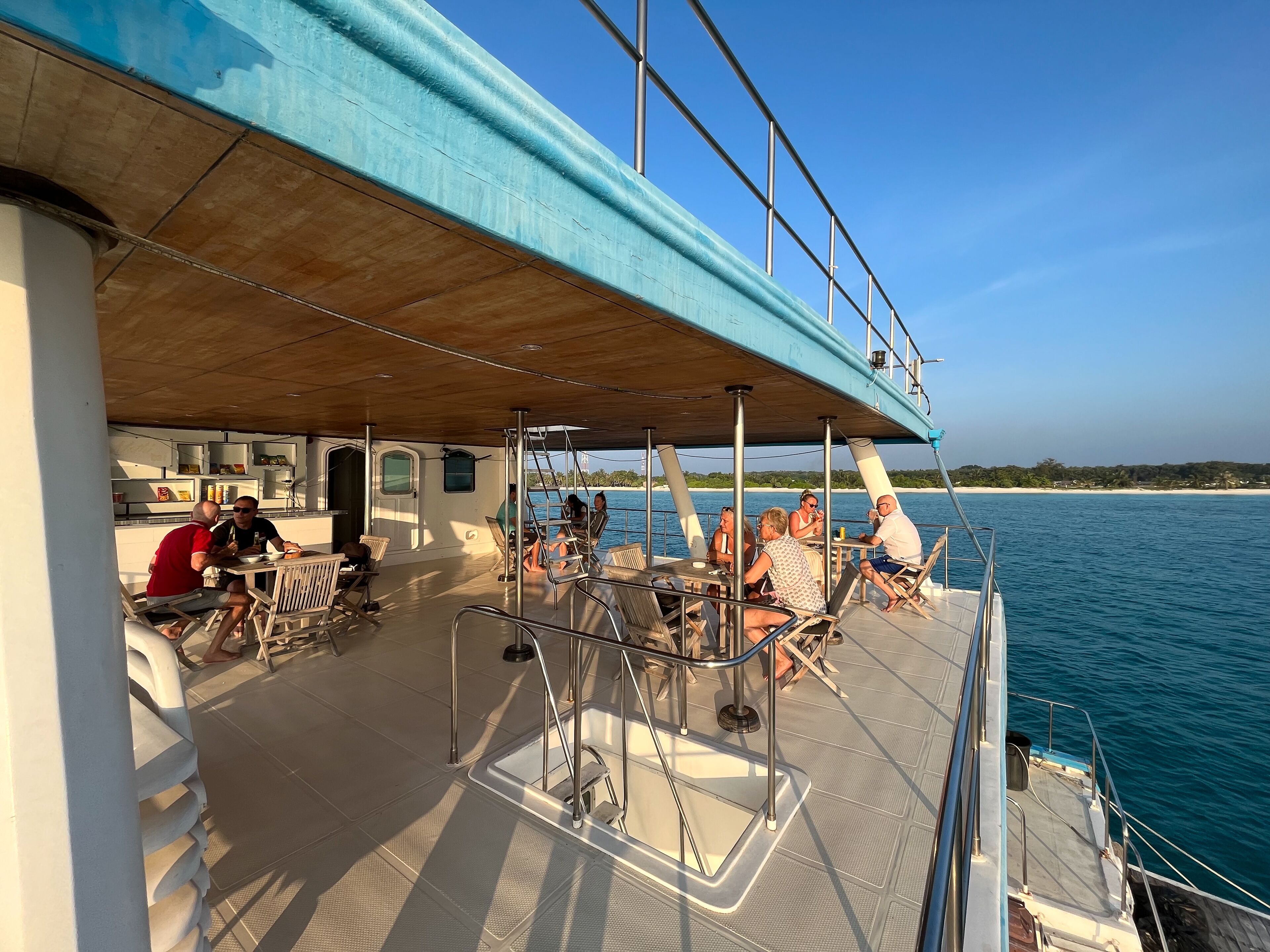 Un espace extérieur abrité sur un bateau, avec des tables et des chaises, offrant une vue sur une étendue d’eau et une plage.