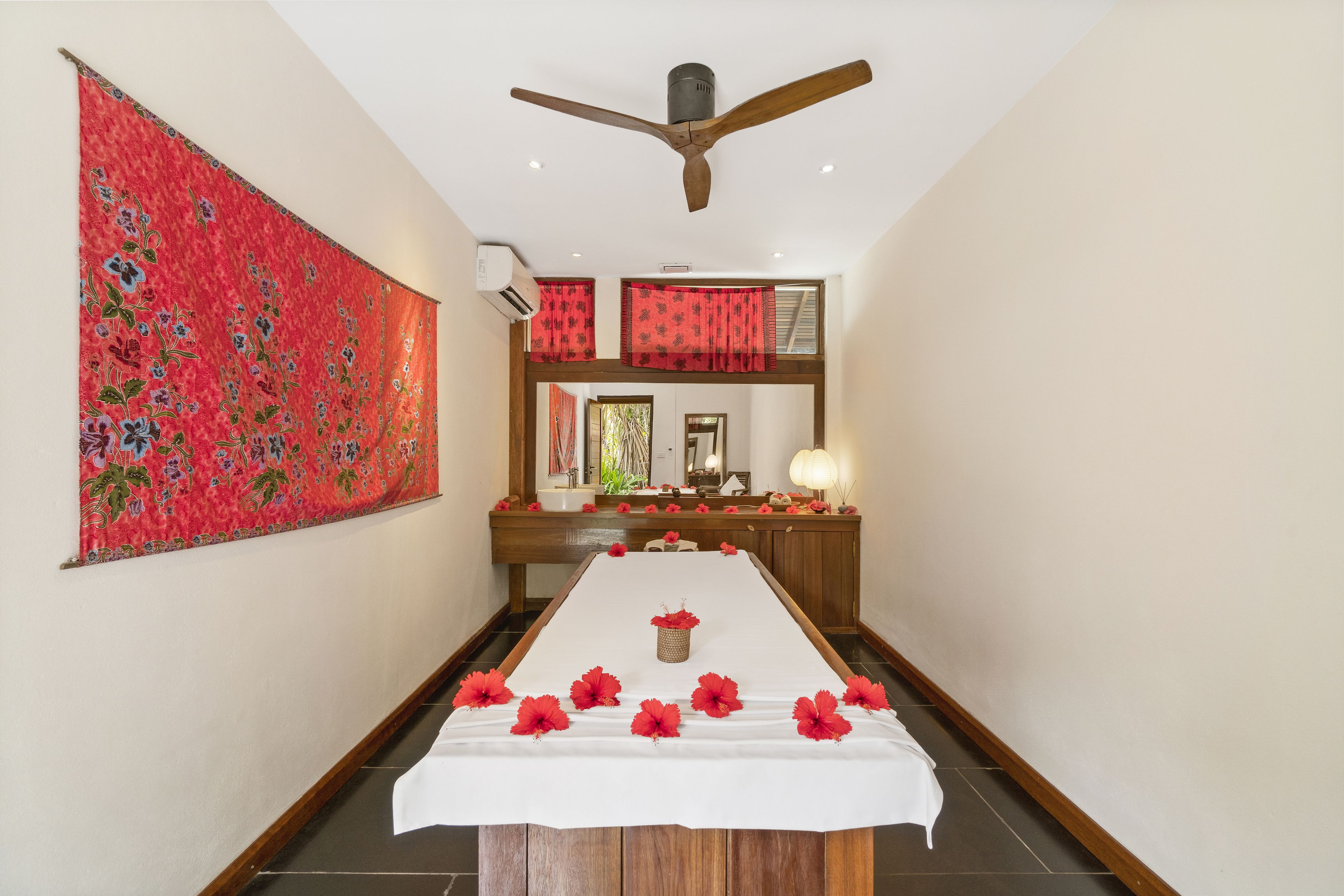 A hotel room with a long dining table, red floral curtains, a ceiling fan, and a large mirror above the table.
