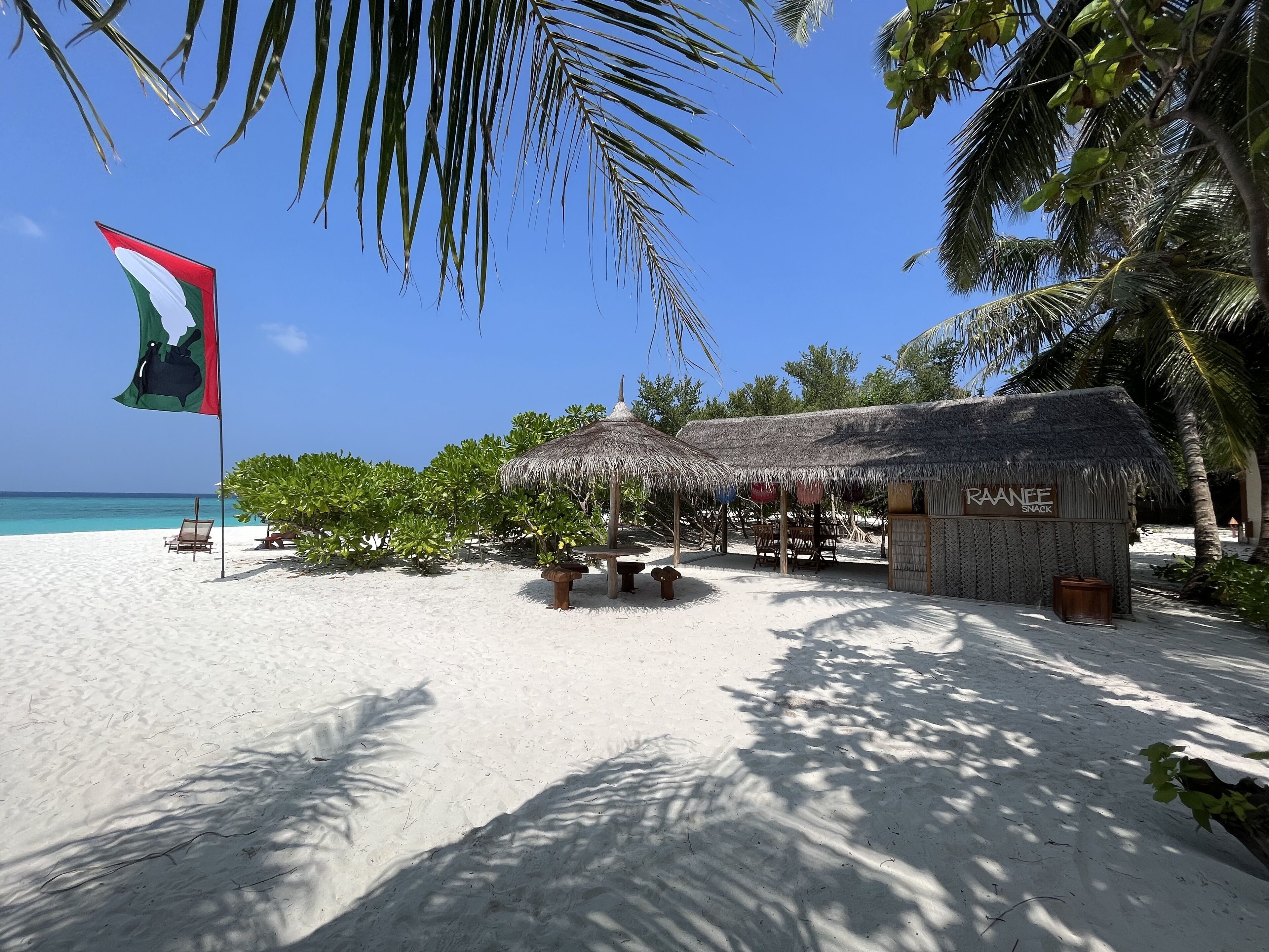A beach with a flag, thatched-roof huts, and a snack bar named "RAANEE".