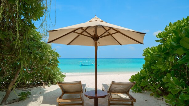 A beach scene with a table and two chairs under a large umbrella, overlooking a clear blue sea and a distant boat. - The Barefoot Eco Hotel (Hanimaadhoo)