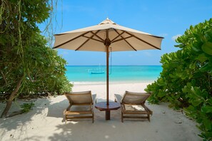A beach scene with a table and two chairs under a large umbrella, overlooking a clear blue sea and a distant boat. - The Barefoot Eco Hotel (Hanimaadhoo)