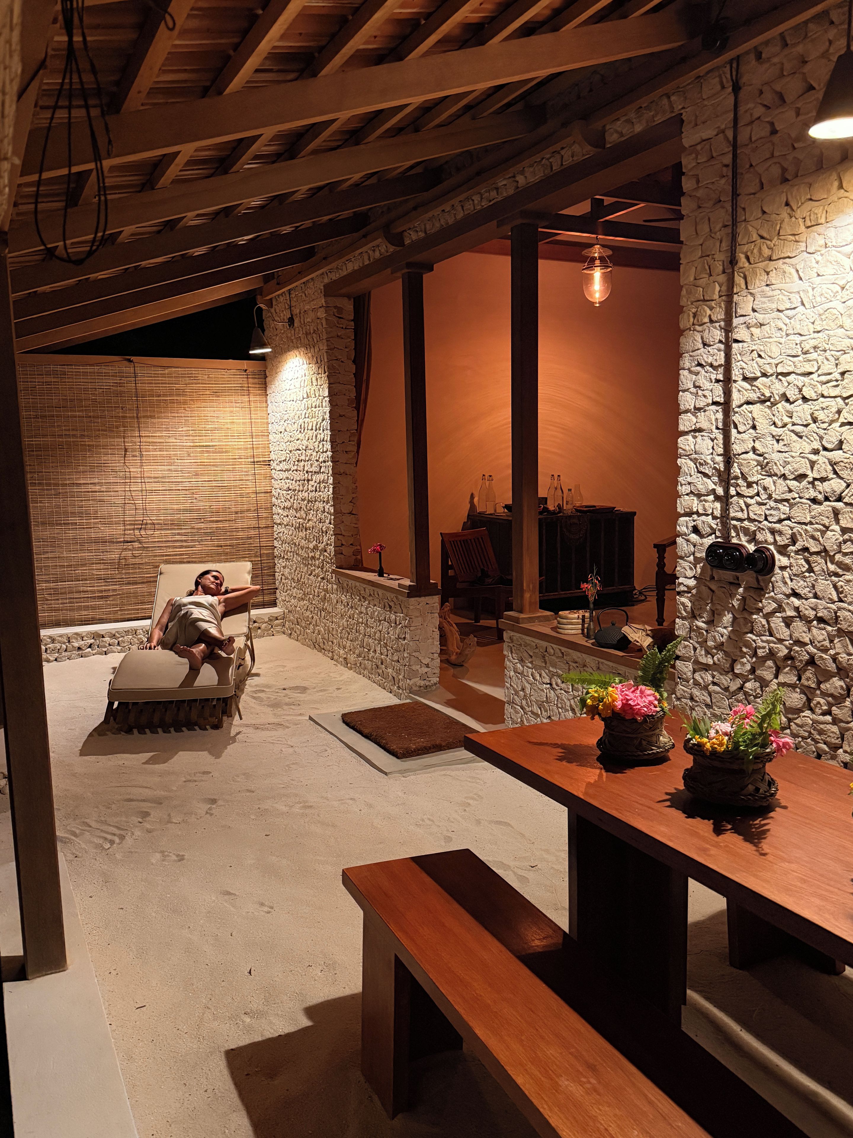 Un interior acogedor con pared de piedra, vigas de madera, un banco y una mesa con flores.