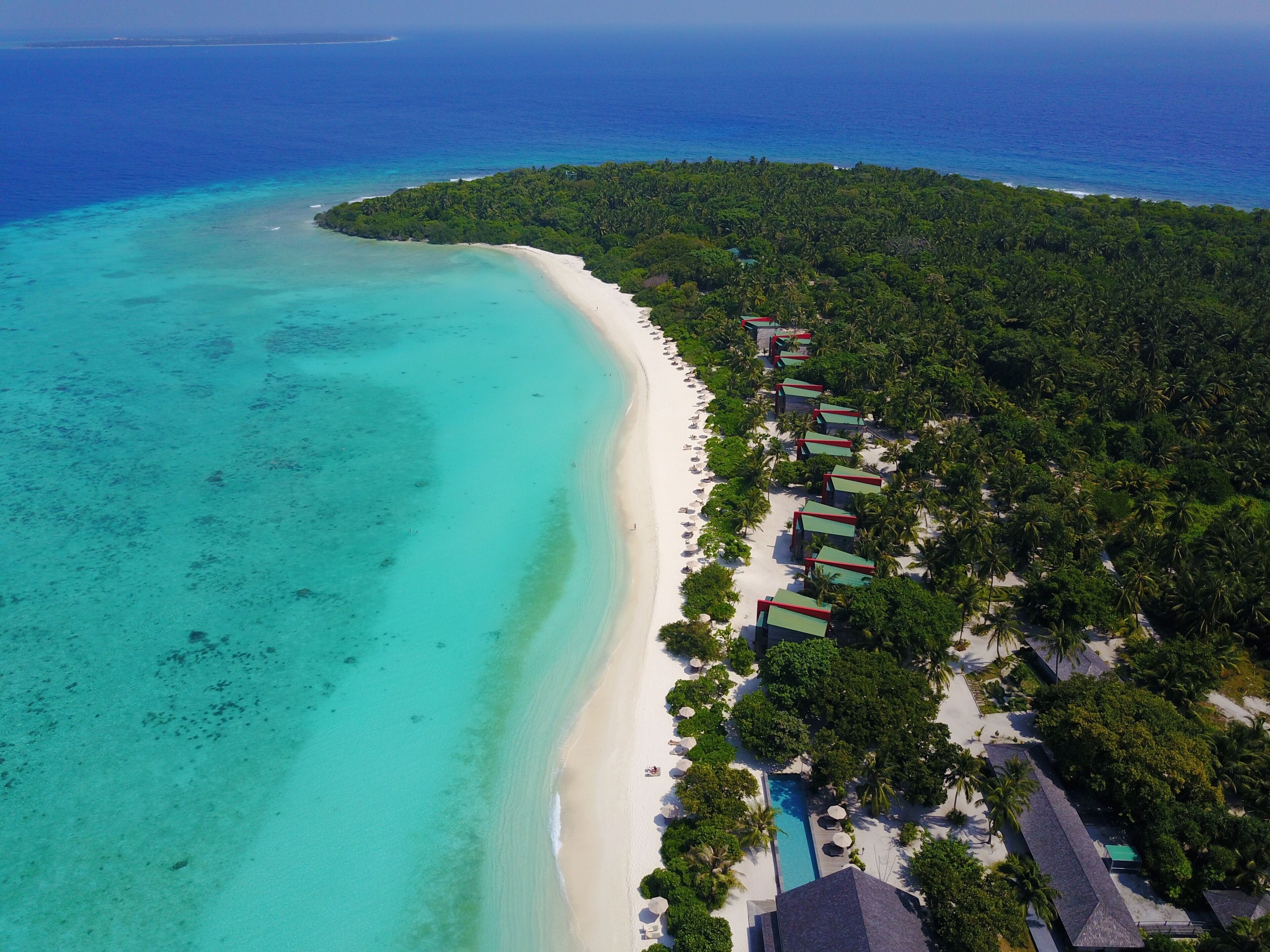 Vue aérienne d’un complexe hôtelier tropical situé sur une plage, avec une longue étendue de sable blanc, des eaux turquoise et limpides, et une végétation luxuriante.