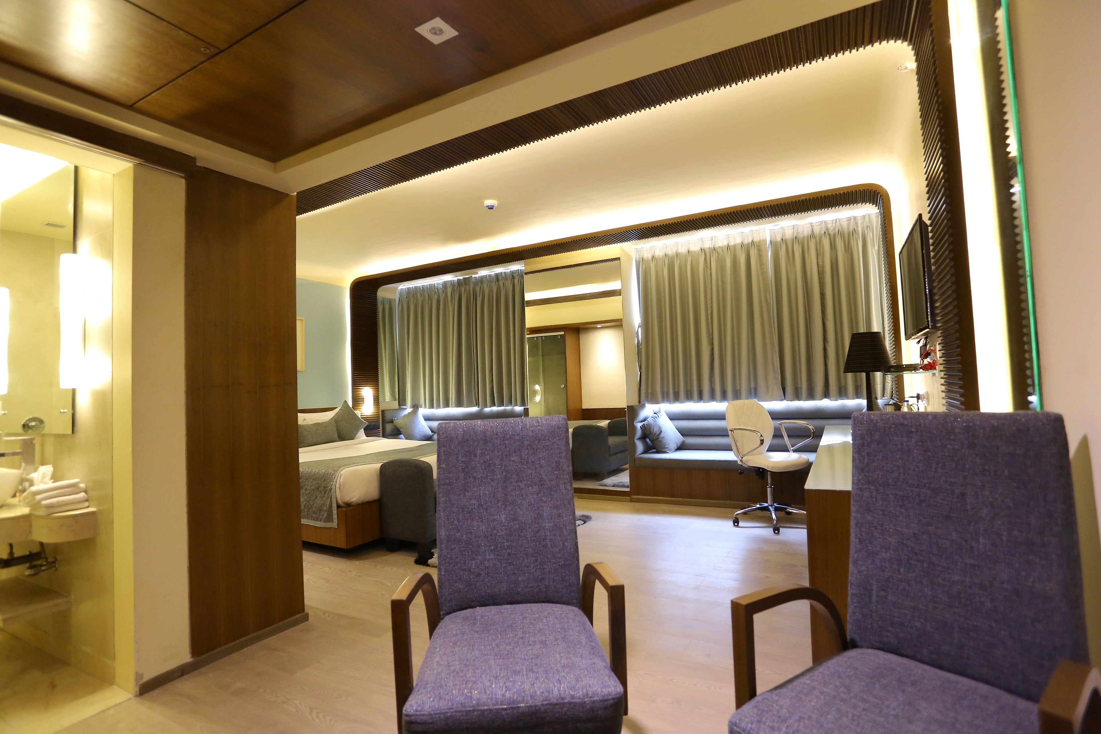 A modern hotel room with a large bed, a seating area with a sofa and armchairs, a desk with a lamp, and a large mirror.