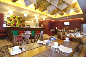 A hotel dining area with tables set for breakfast, a reception desk, and a TV screen on the wall.