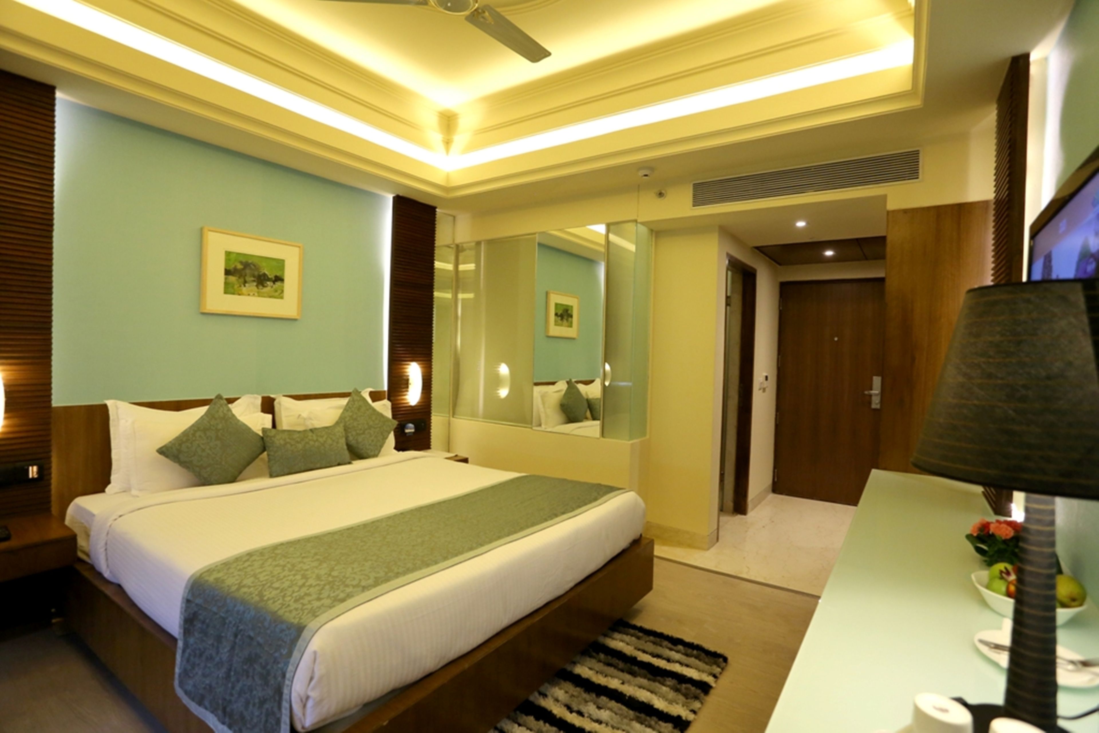 A hotel room with a large bed, bedside tables, a desk, a chair, a lamp, a mirror, and a door.