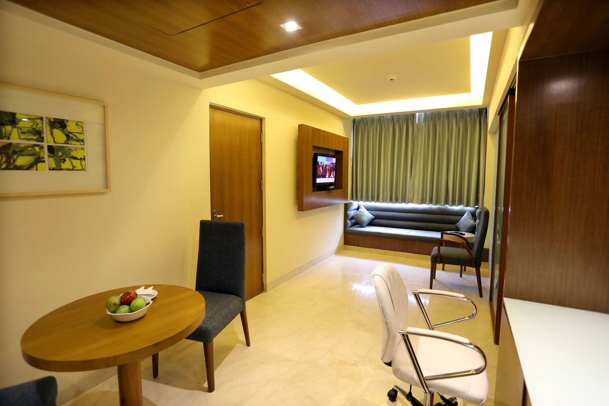 A modern hotel room with a wooden ceiling, a dining table with a bowl of fruit, a TV mounted on the wall, a sofa, and a chair.