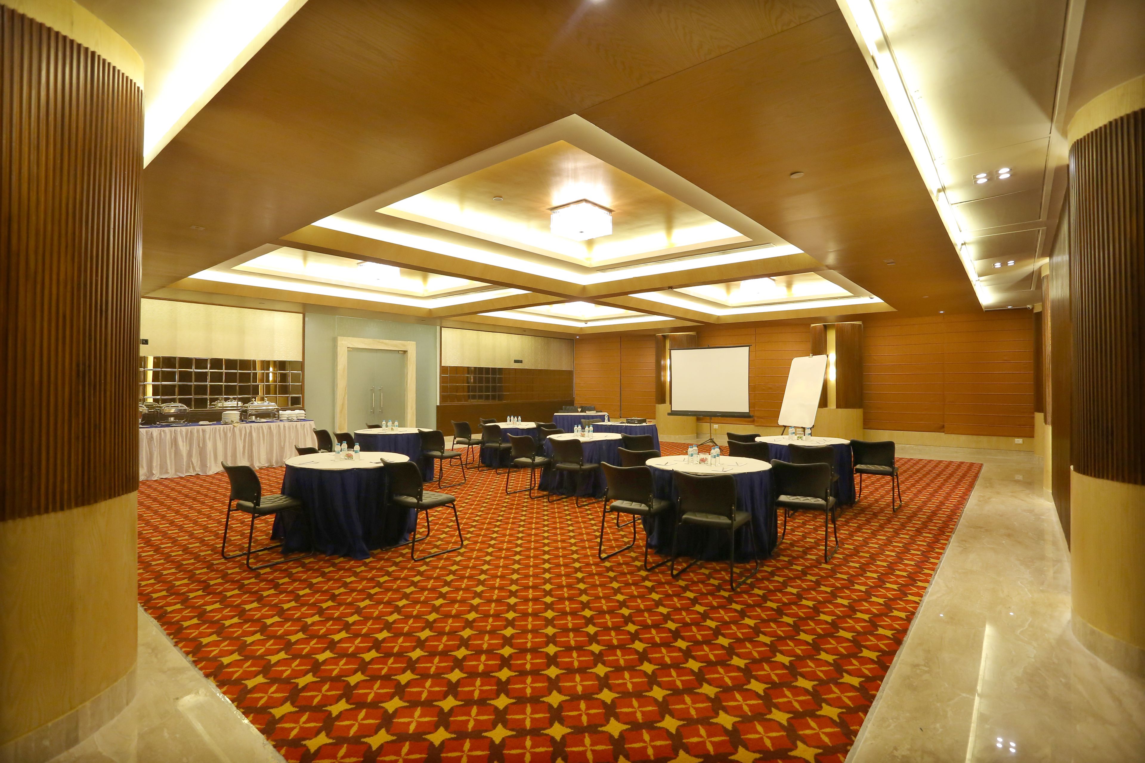 A conference room with a patterned carpet, round tables, and chairs, equipped with a projector screen and a whiteboard.