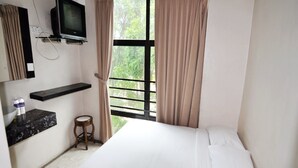 A hotel room with a bed, a TV mounted on the wall, a small stool, and a window with curtains.