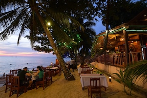 Un área de comedor al aire libre con mesas y sillas, palmeras y vista al mar.