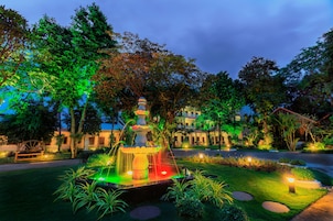 Un jardín bien iluminado con una fuente, rodeado de árboles y un edificio al fondo.