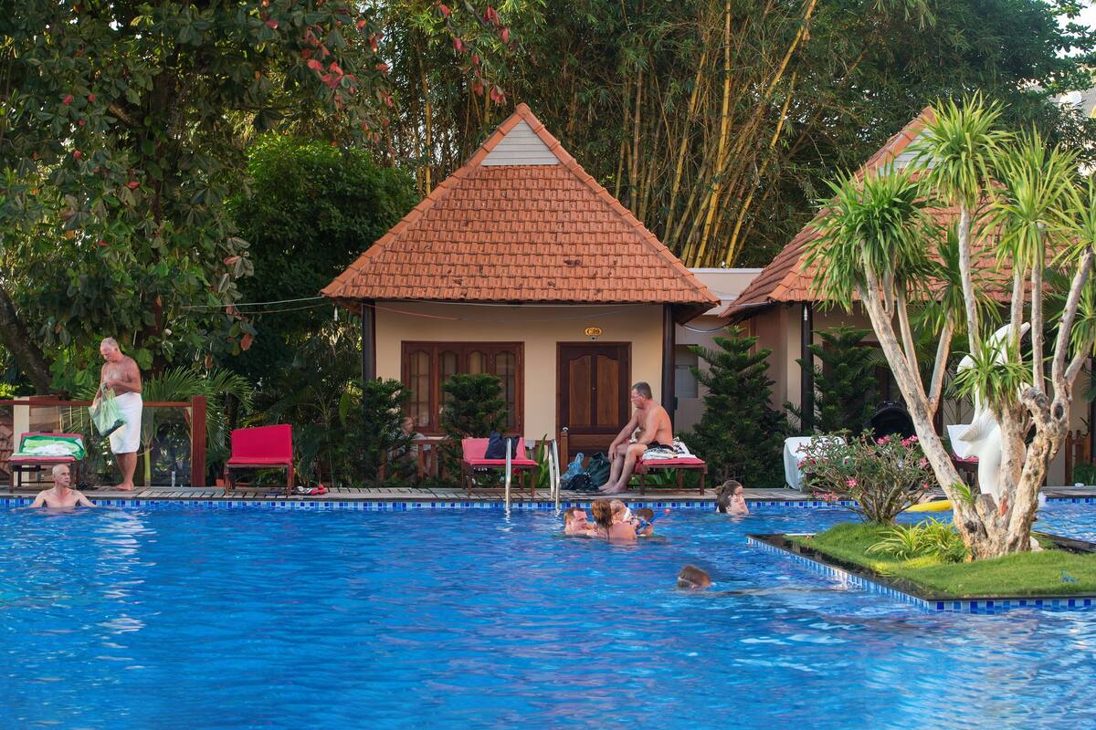 A swimming pool with people in it, surrounded by greenery and a building with a tiled roof.