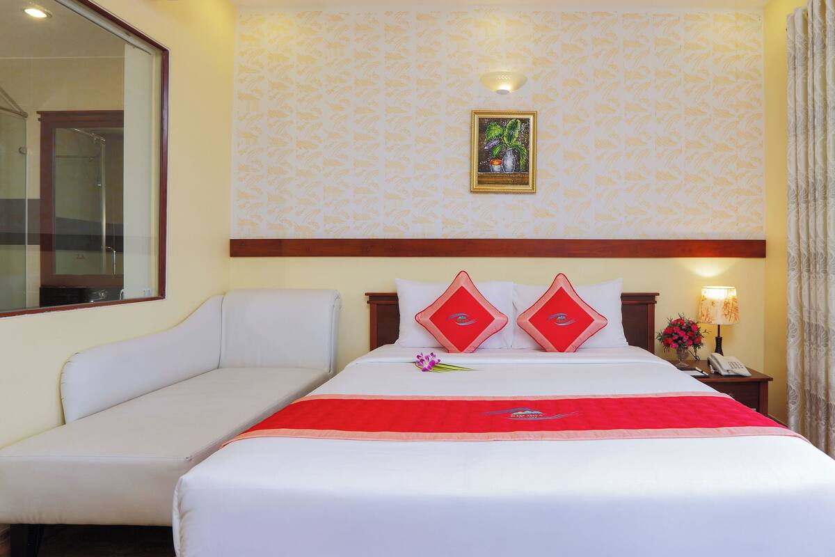 A hotel room with a bed, a sofa, a nightstand, a lamp, and a view of the ocean.