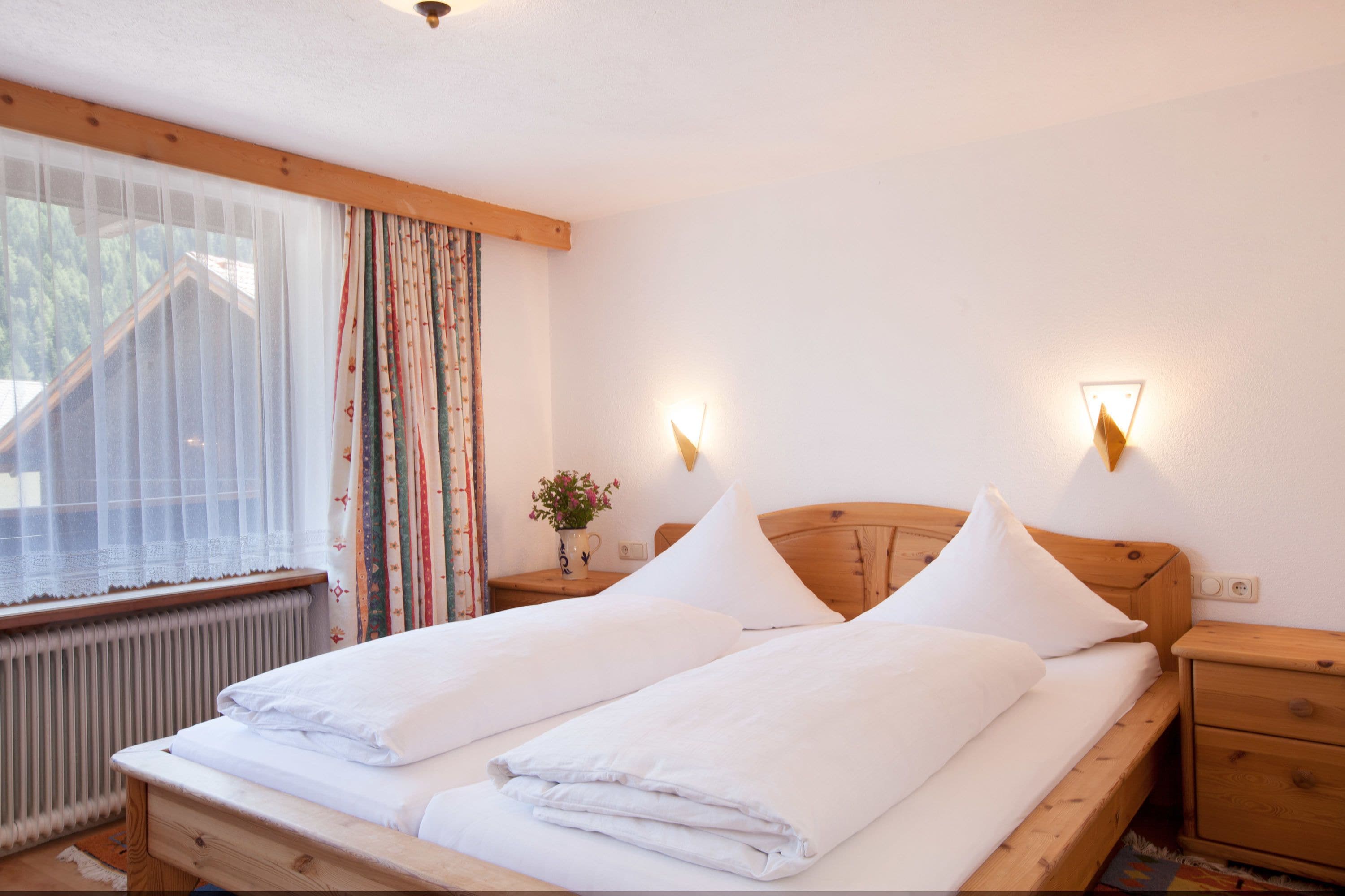 A double bed with white linens, a wooden headboard, two bedside tables with lamps, a window with sheer curtains, and a radiator.