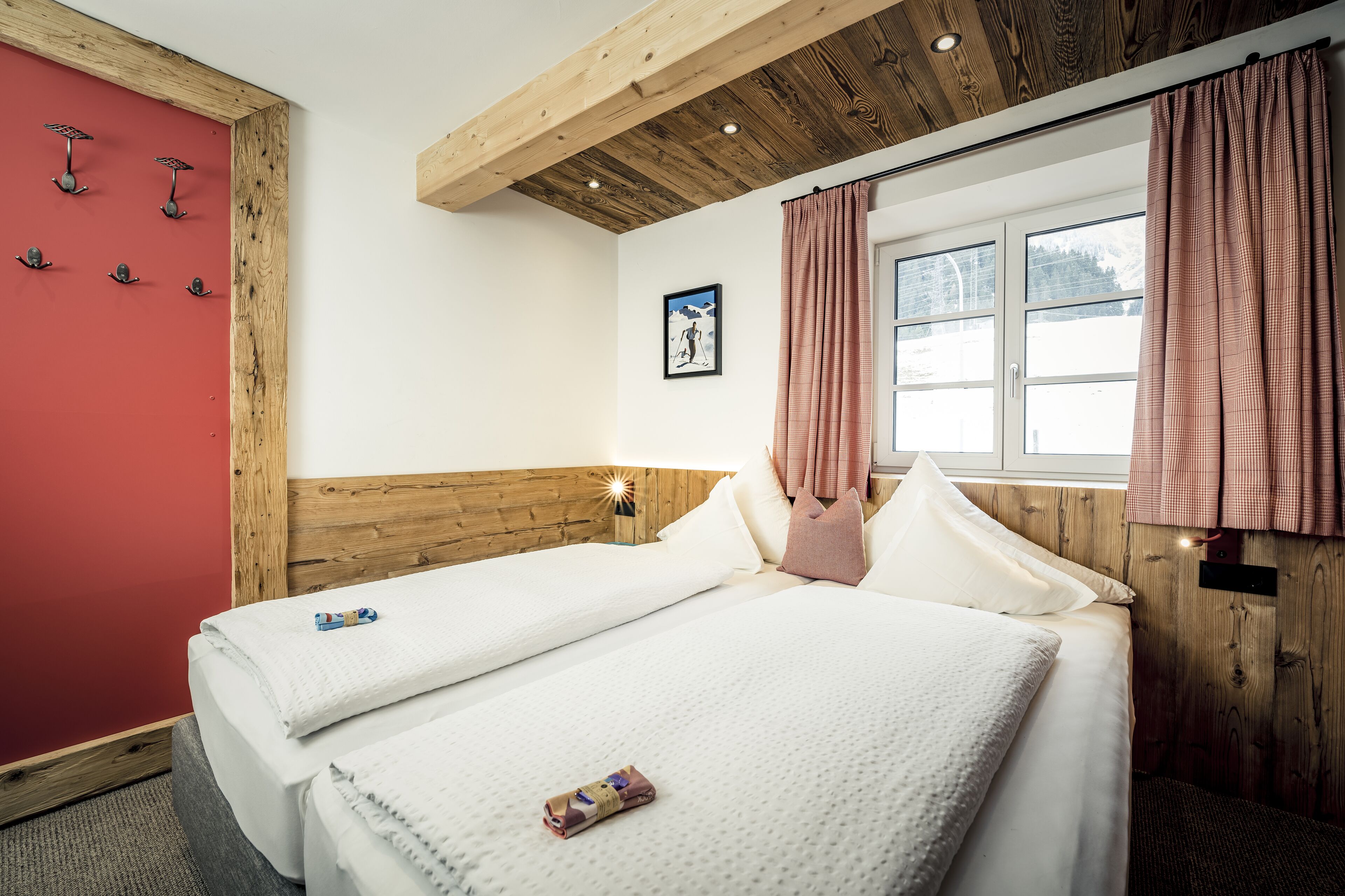 Habitación de hotel con dos camas, vigas de madera en el techo, una pared roja y una ventana con cortinas.
