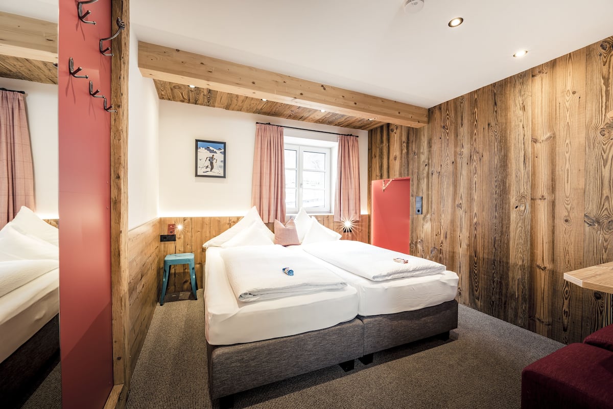 Una cama doble con cabecera de madera, un taburete pequeño y una ventana con cortinas.