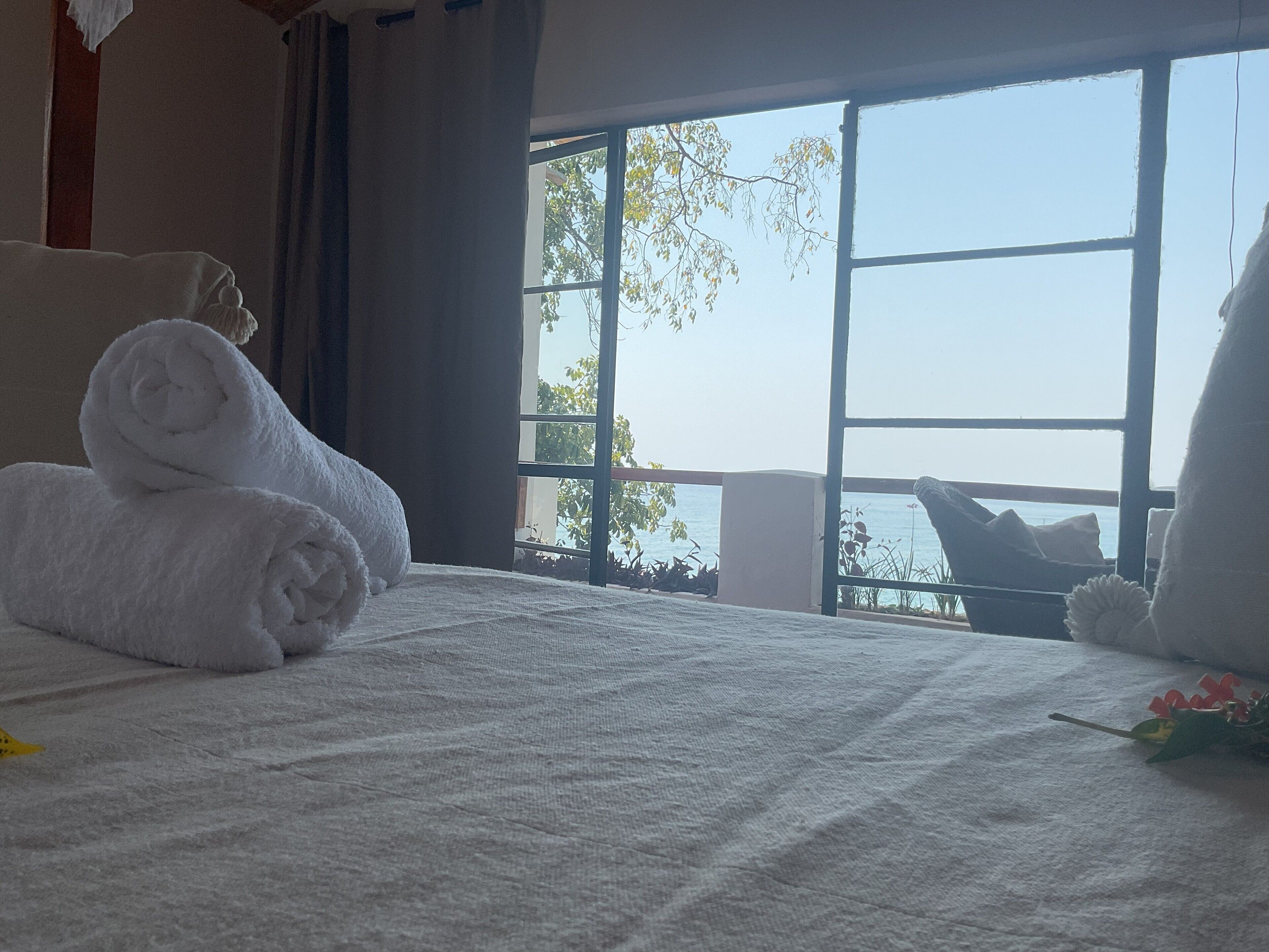 A hotel room with a bed, a window with a view of the sea, and a balcony with a chair.