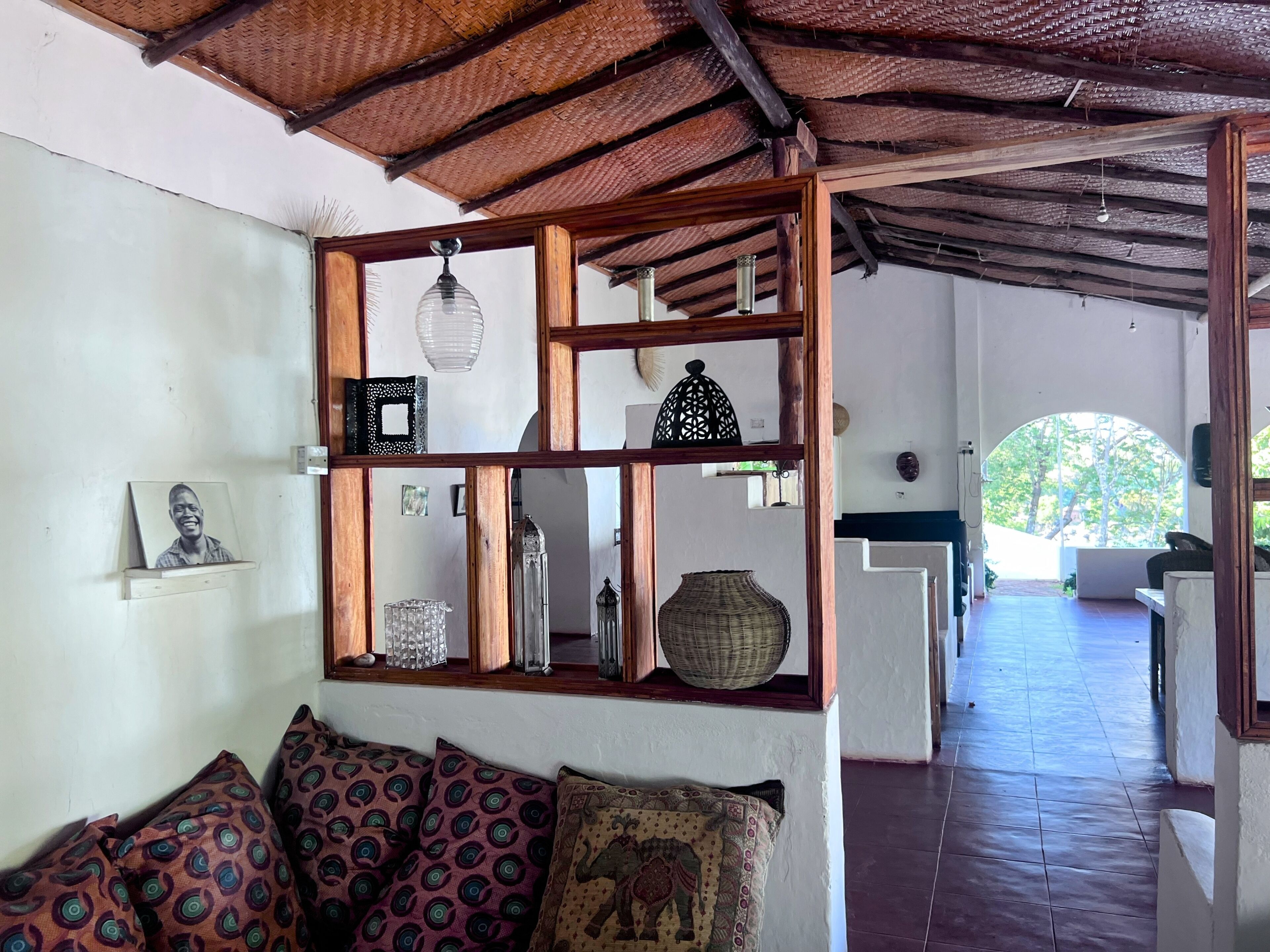 A room with a wooden ceiling, a sofa with patterned cushions, a shelf with decorative items, and a view of an outdoor area through an arched doorway.