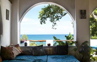 A room with a view of the sea through an arched window, a bed with patterned bedding, and a plant on the windowsill.