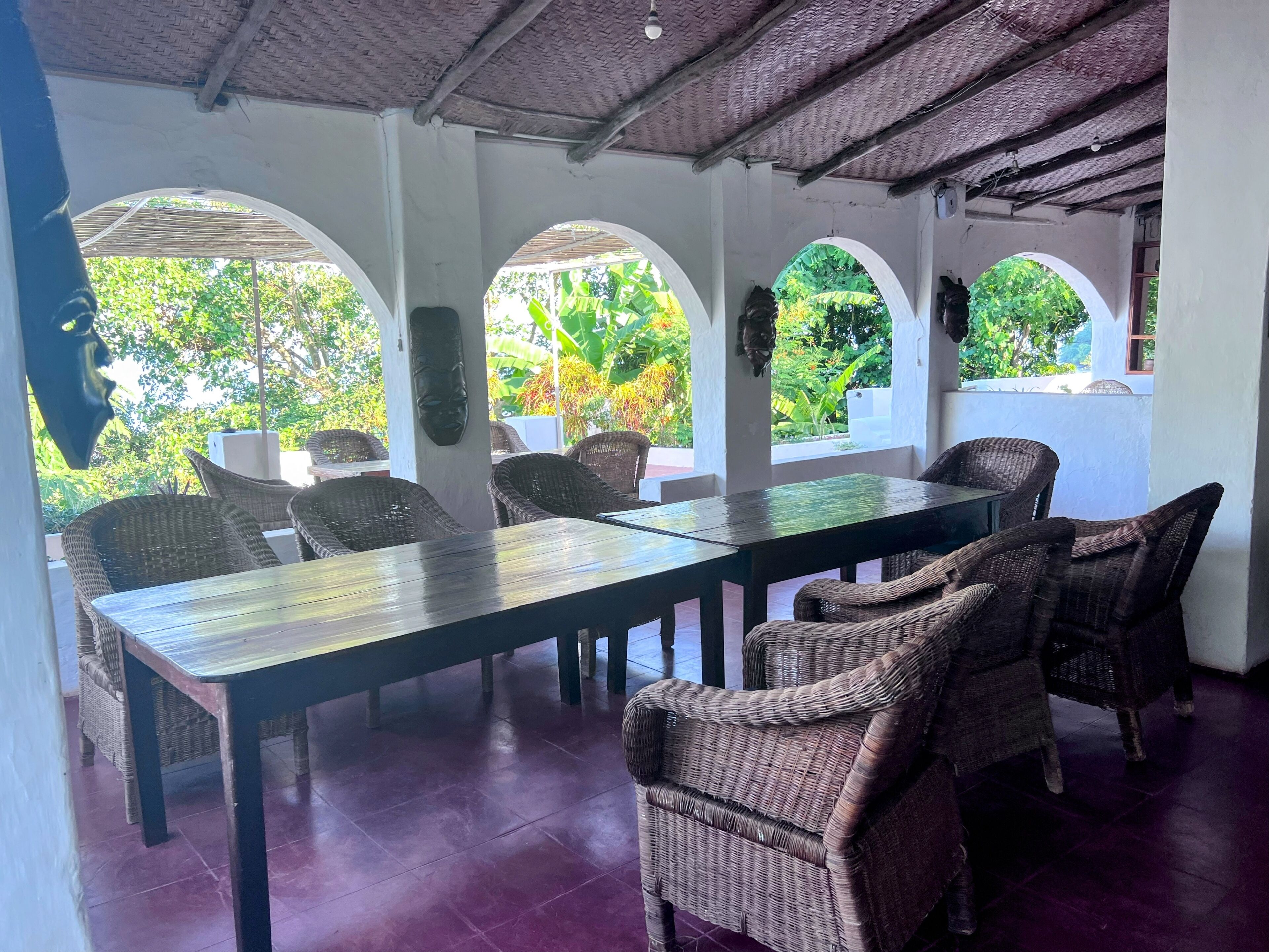 A covered outdoor dining area with wicker furniture and a long table, surrounded by arches with views of greenery.