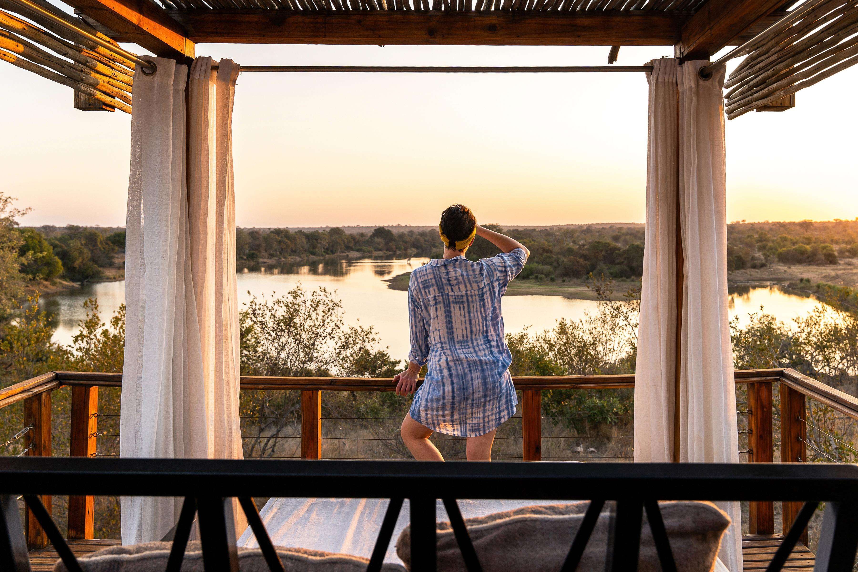 A person standing on a wooden deck overlooking a river with trees and hills in the background.