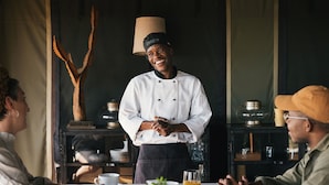 Un chef de pie junto a una mesa con comensales, una lámpara y estantes con utensilios de cocina.