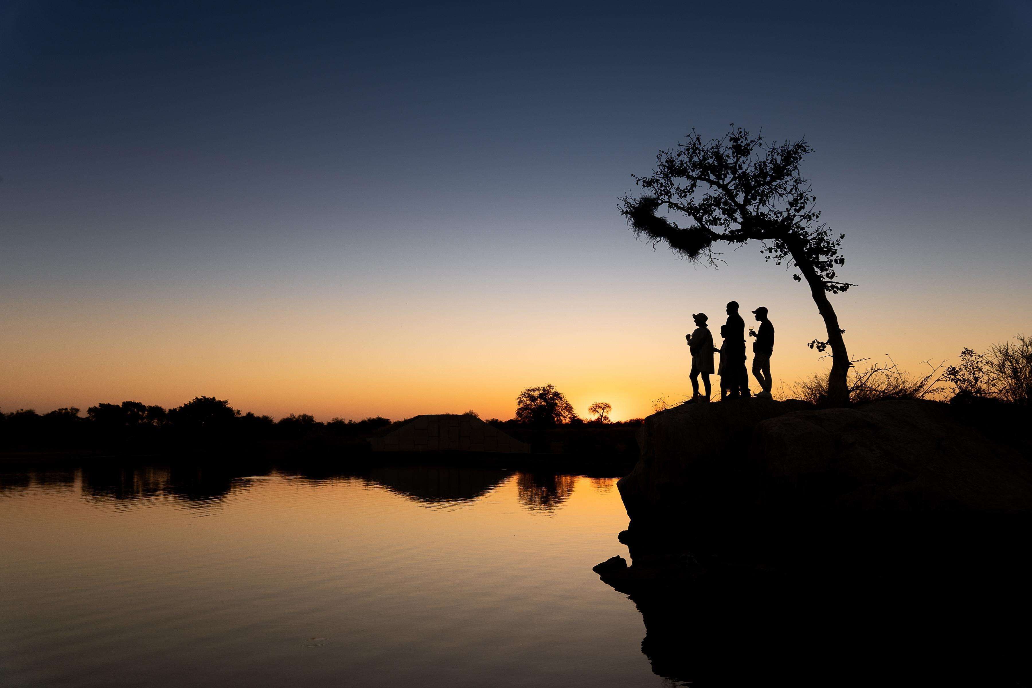 La silhouette de trois personnes se tenant près d’une étendue d’eau calme, au coucher du soleil.