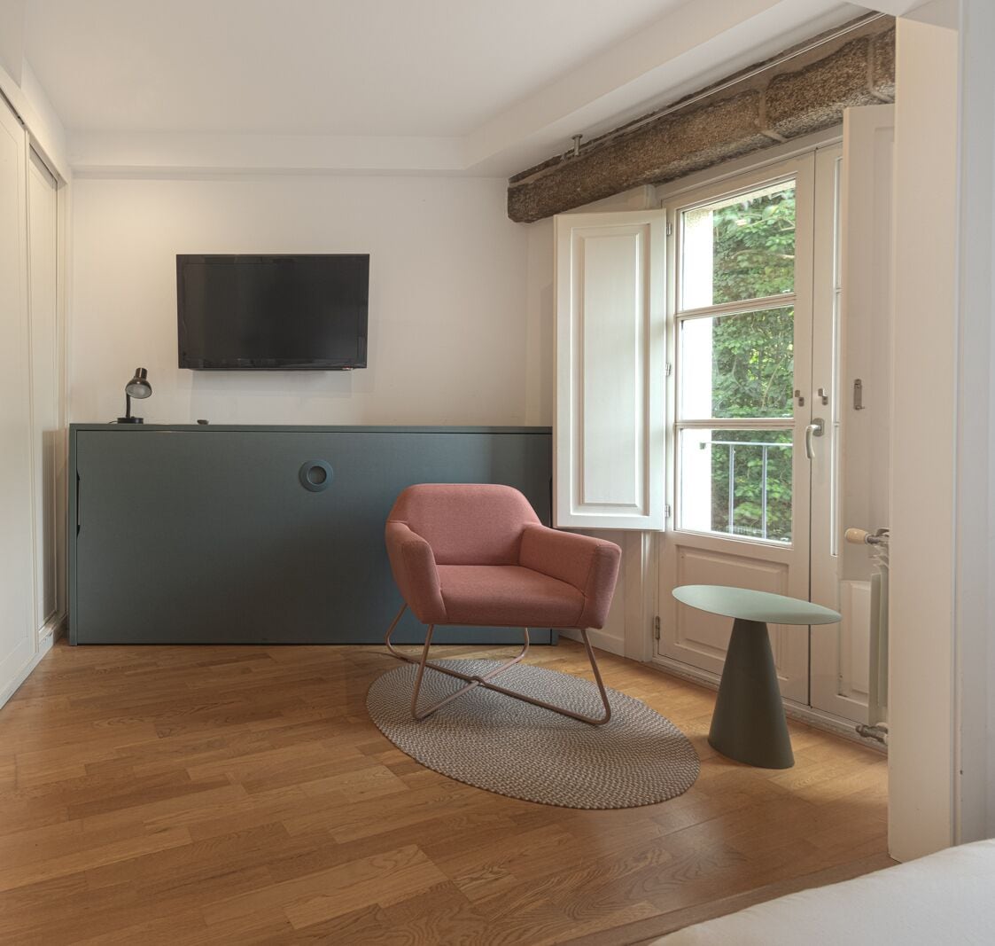 A room with a wooden floor, a pink chair, a small round table, a television on the wall, and a window with a view of greenery.