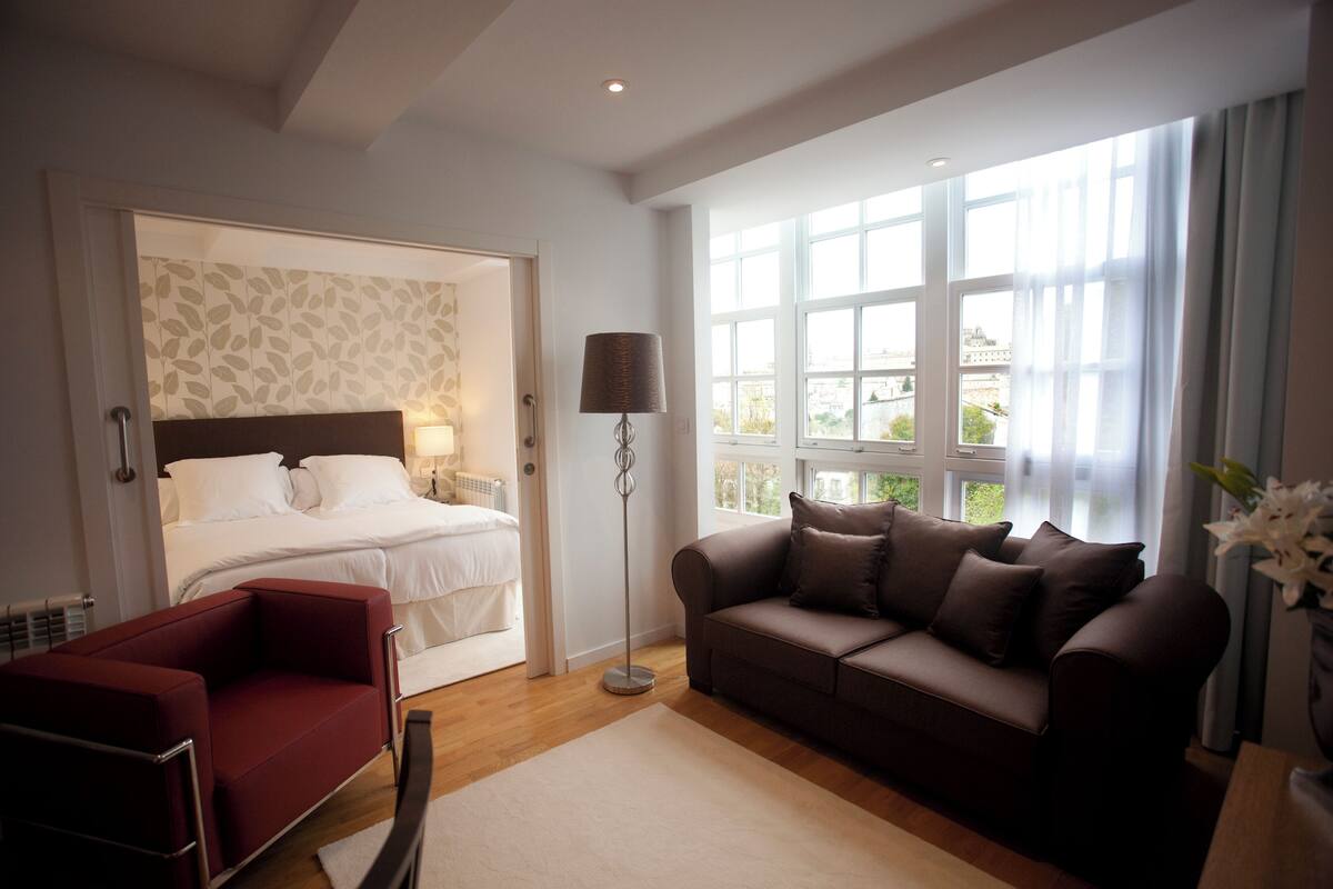 A hotel room with a bed, a red armchair, a brown sofa, a floor lamp, and a large window with curtains.