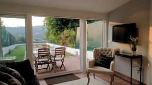 Un salón con sofá, una silla, televisor y un ventanal corredero que da a un patio exterior con mesa y sillas.