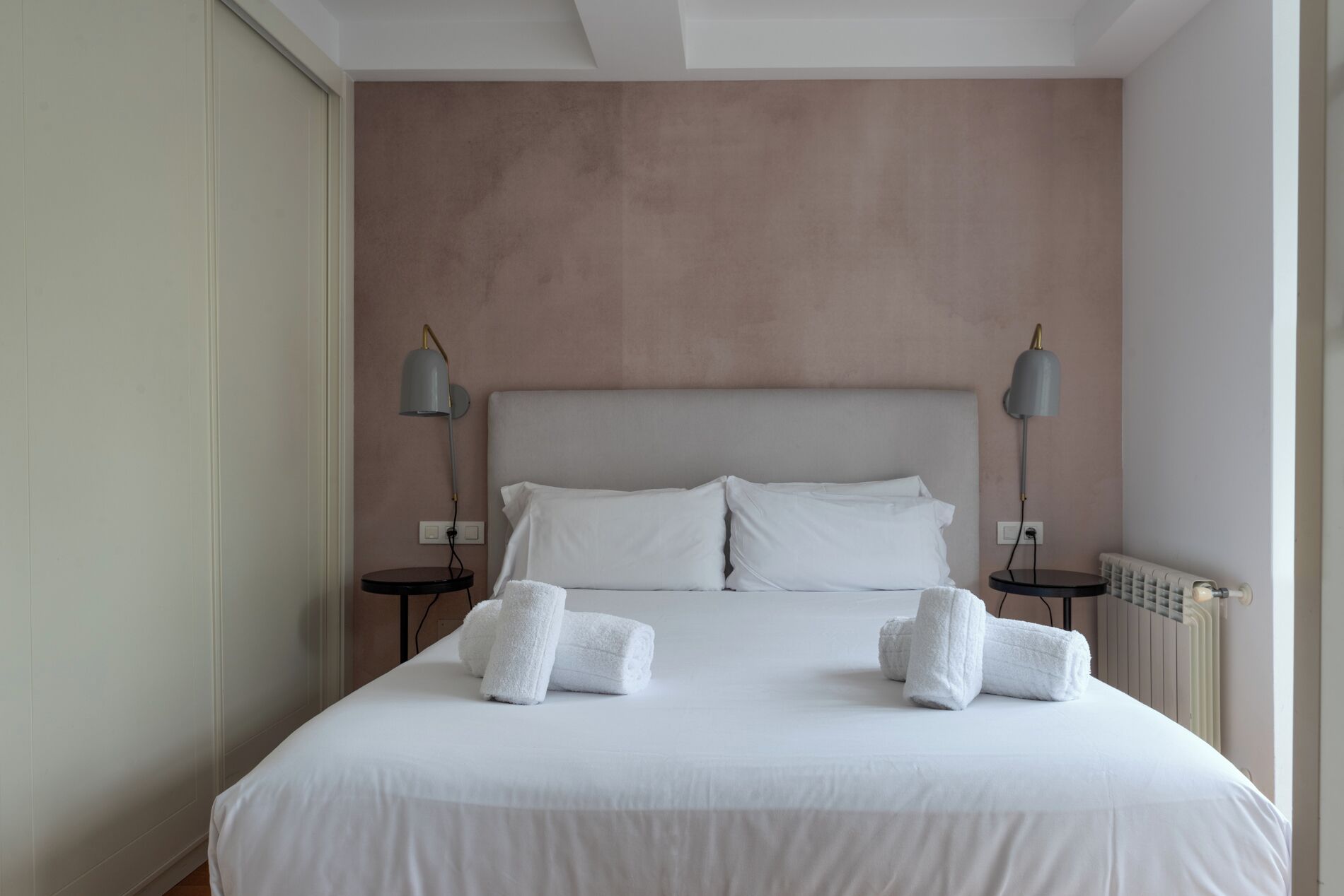 A neatly made bed with white linens and two rolled towels, two bedside lamps, and a radiator on the wall.