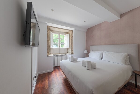 Un dormitorio con una cama grande, un televisor montado en la pared, una ventana con vistas a zonas verdes y un radiador debajo de la ventana.