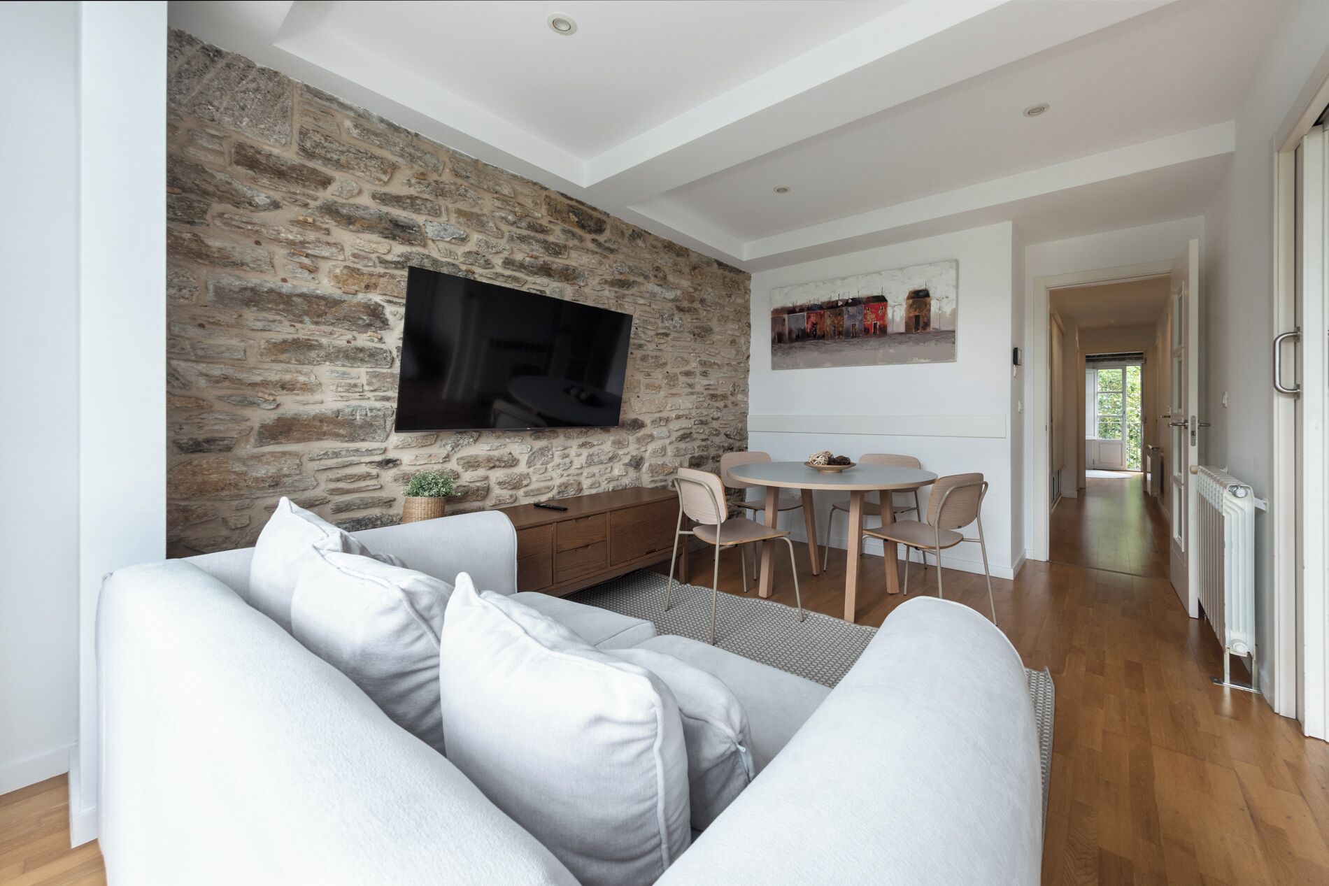 A modern living room with a stone wall, a flat-screen TV, a grey sofa, and a dining area with a round table and chairs.