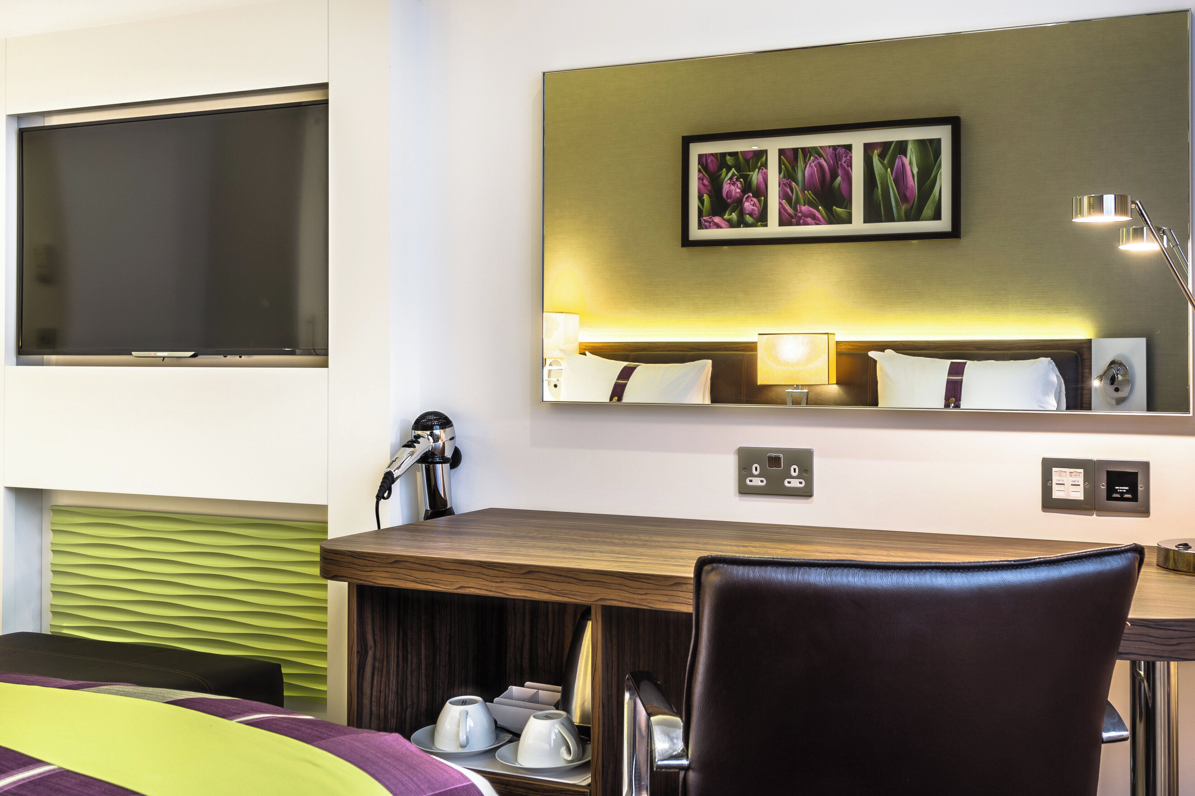 A hotel room with a bed, a desk with a chair, a television, a mirror, and a wall-mounted power outlet.
