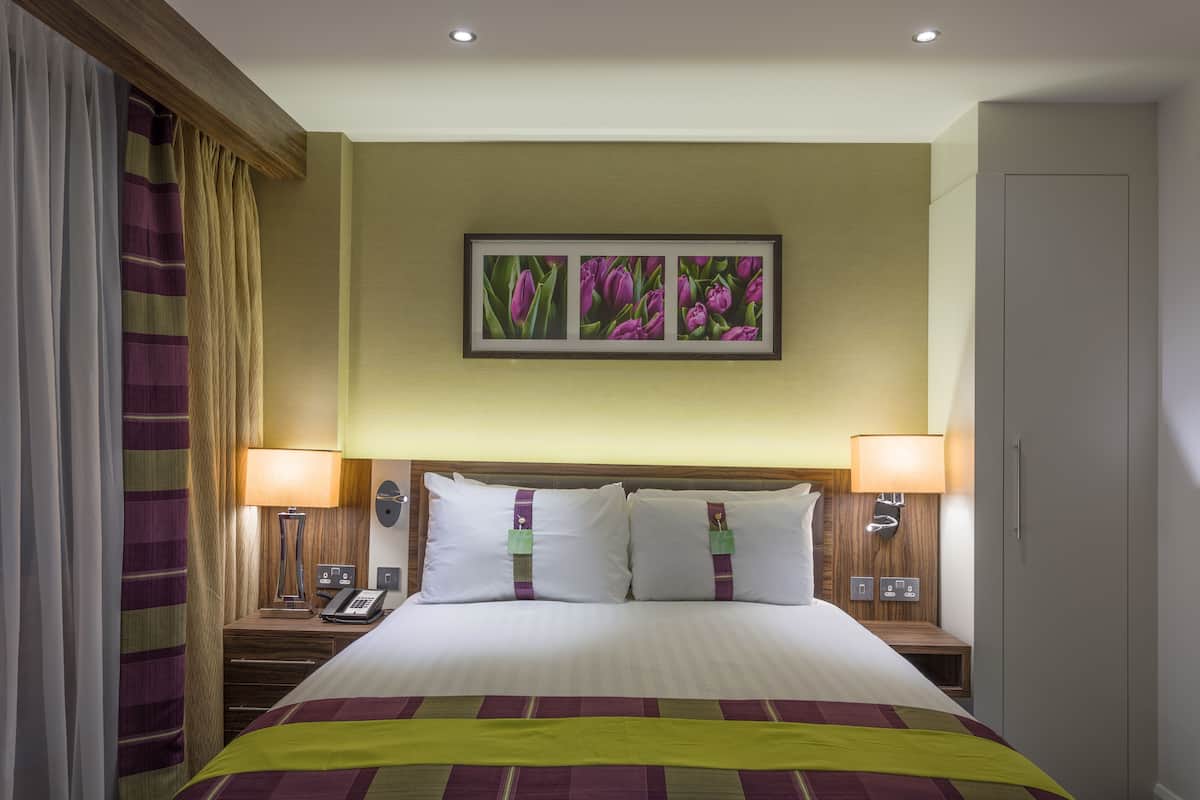 A hotel room with a large bed, two bedside tables with lamps, a wall-mounted telephone, and a framed picture above the bed.