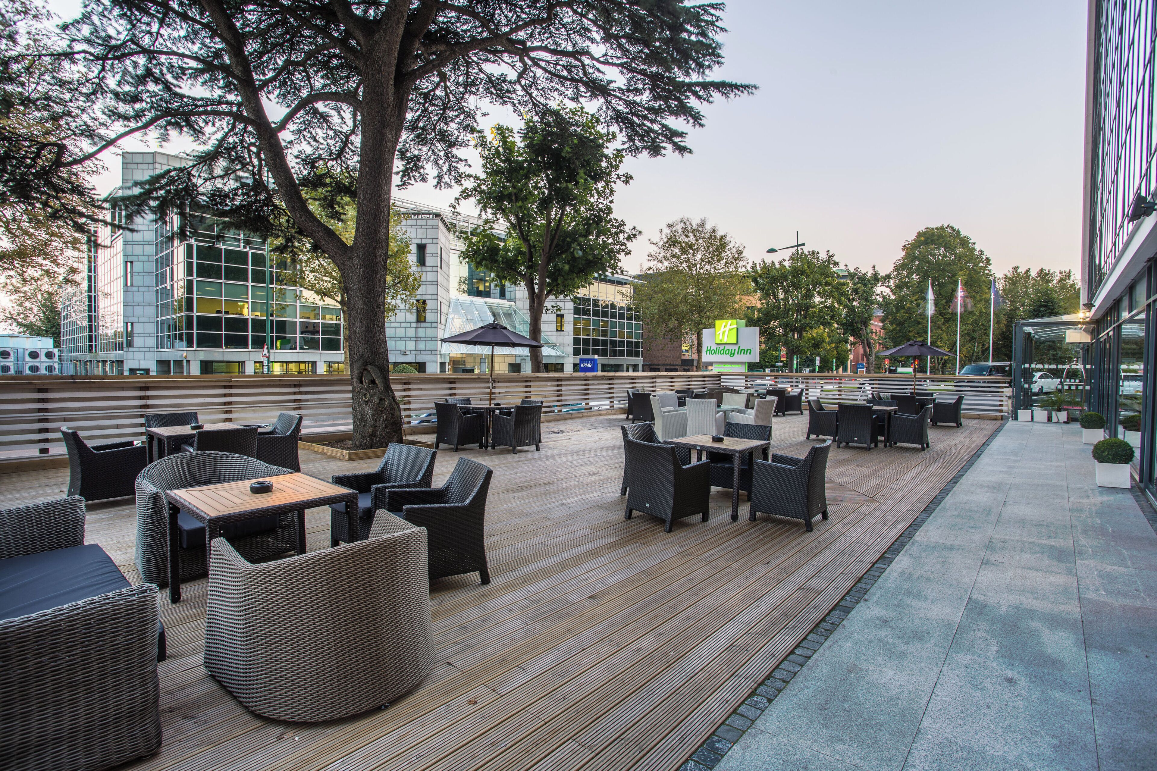 Un espacio de estar al aire libre con mobiliario de mimbre y una terraza de madera, con vistas a un edificio moderno de fachada de vidrio.