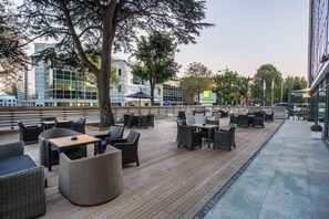 Un espacio de estar al aire libre con mobiliario de mimbre y una terraza de madera, con vistas a un edificio moderno de fachada de vidrio.