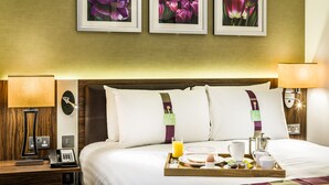 A hotel room with a neatly made bed, a bedside table with a lamp, a tray with breakfast items, and three framed floral pictures on the wall.