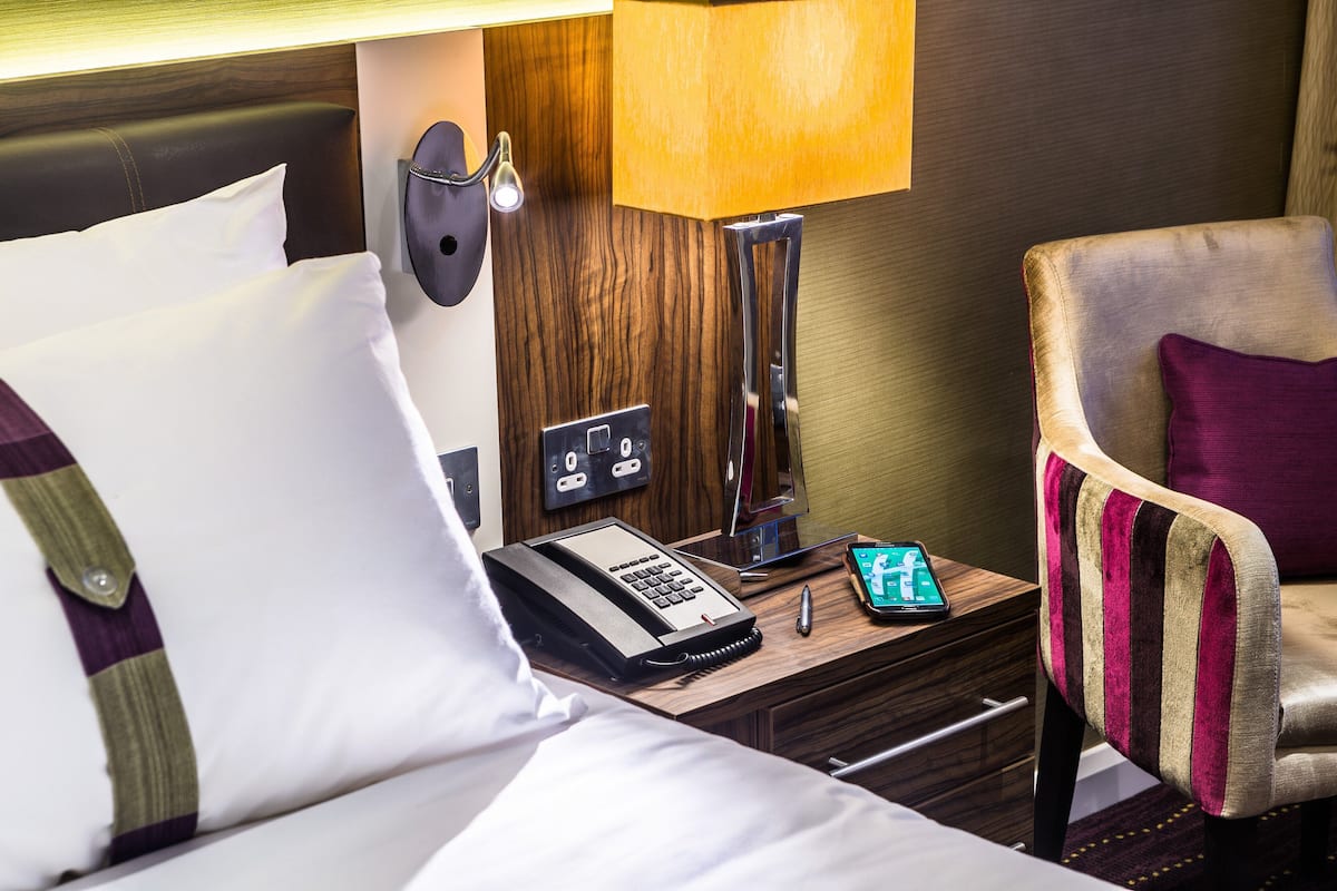 A hotel room with a bed, a nightstand with a lamp, a phone, and a chair.