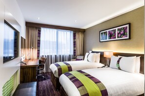 A hotel room with two beds, a desk, a chair, a window with curtains, and a framed picture on the wall. - Holiday Inn London - Watford Junction by IHG (Watford)