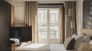 Habitación de hotel con cama, televisor, ventana con cortinas y vistas a edificios.