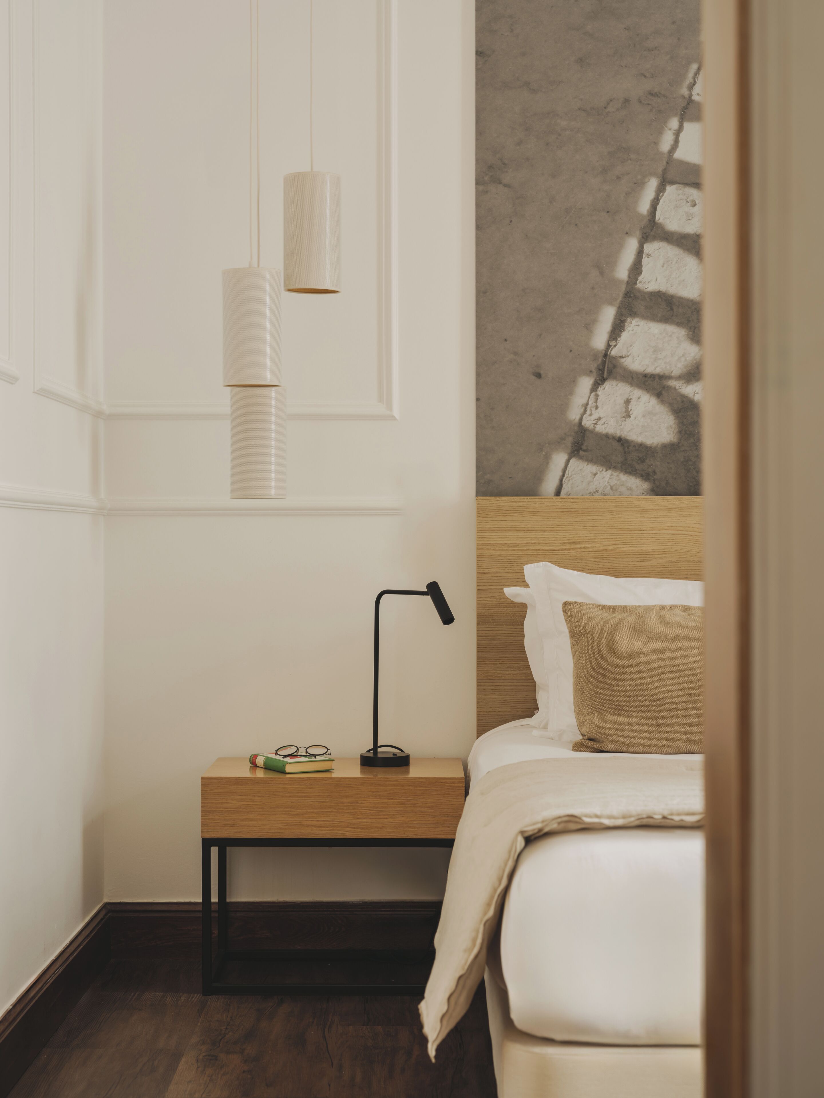 A neatly arranged bedroom with a wooden nightstand, a black desk lamp, a pair of glasses, and a book. The bed has a white headboard and a beige pillow.