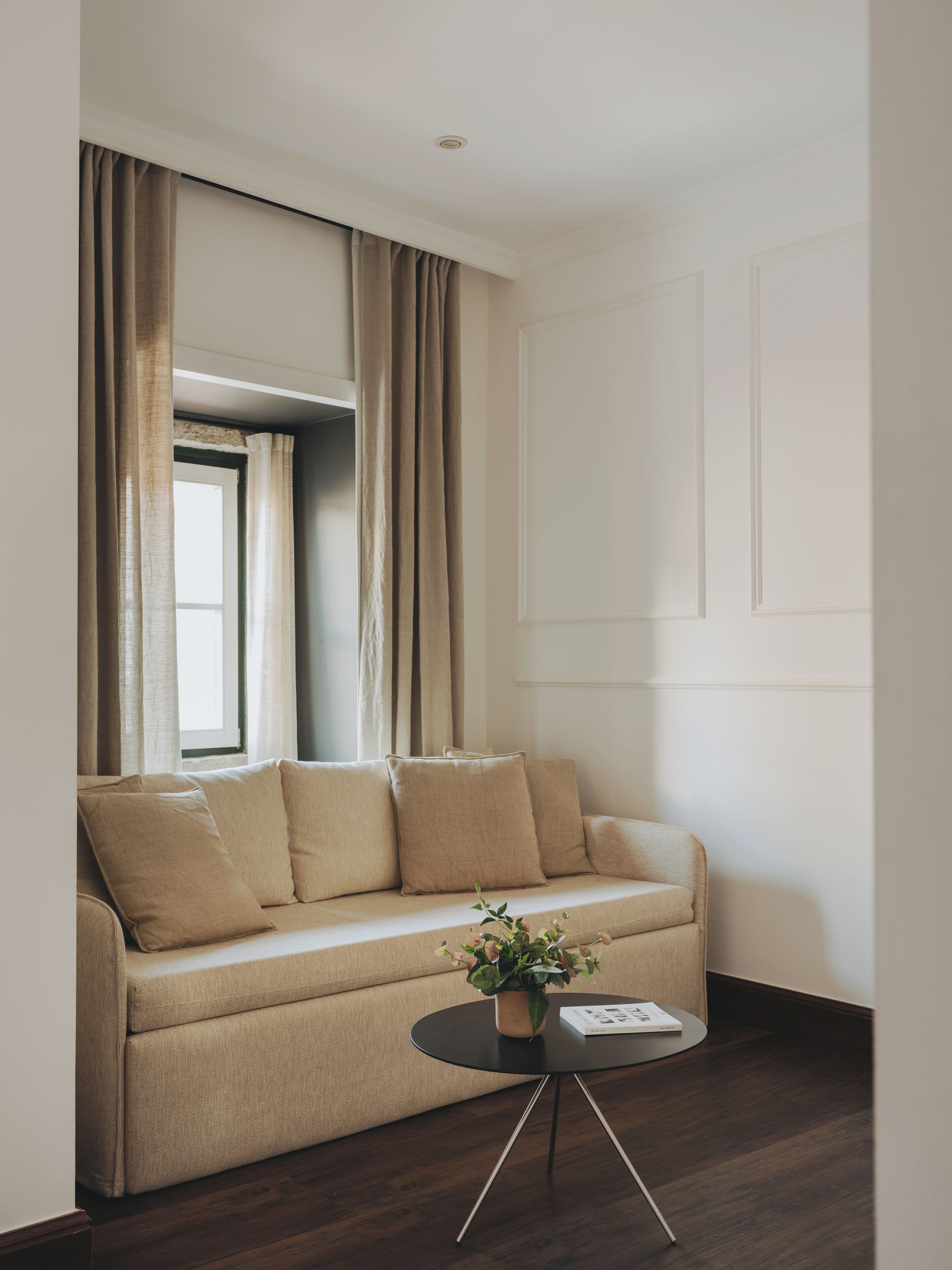 A modern living room with a beige sofa, a small round table with a plant, and a window with curtains.