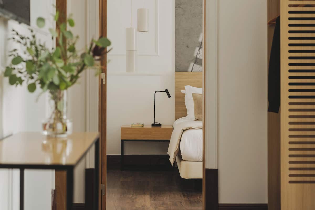 Een moderne hotelkamer met een houten vloer, een nachtkastje met een lamp, een bed met een wit hoofdeinde en een houten garderobe.