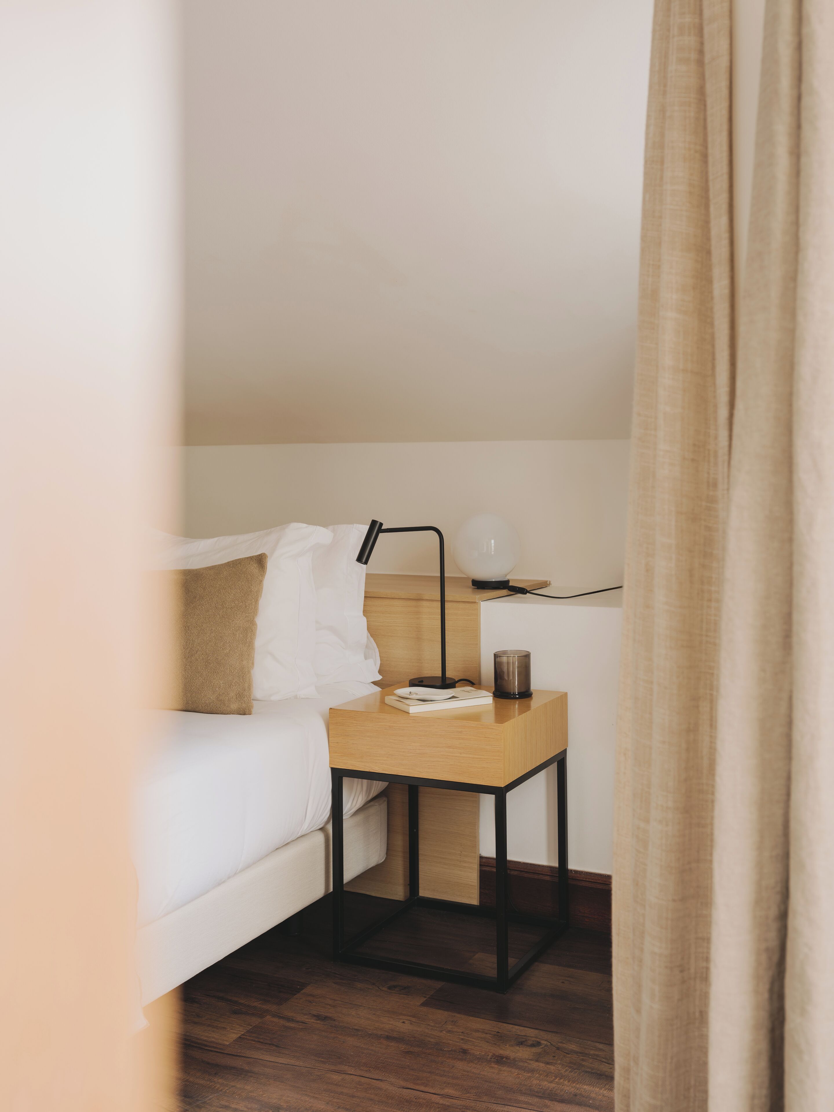 A neatly arranged bedroom with a bed, bedside table, lamp, and a small bedside table with a book and a candle.
