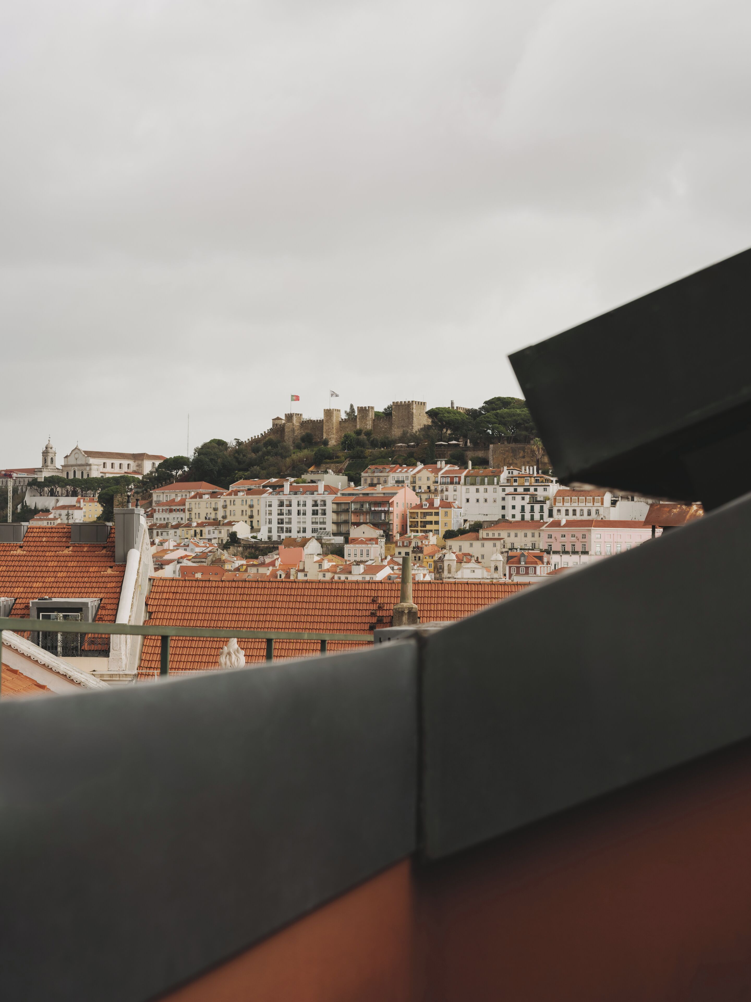 Un paisaje urbano con una estructura imponente similar a un castillo en una colina, rodeado de edificios residenciales con techos de tejas rojas.