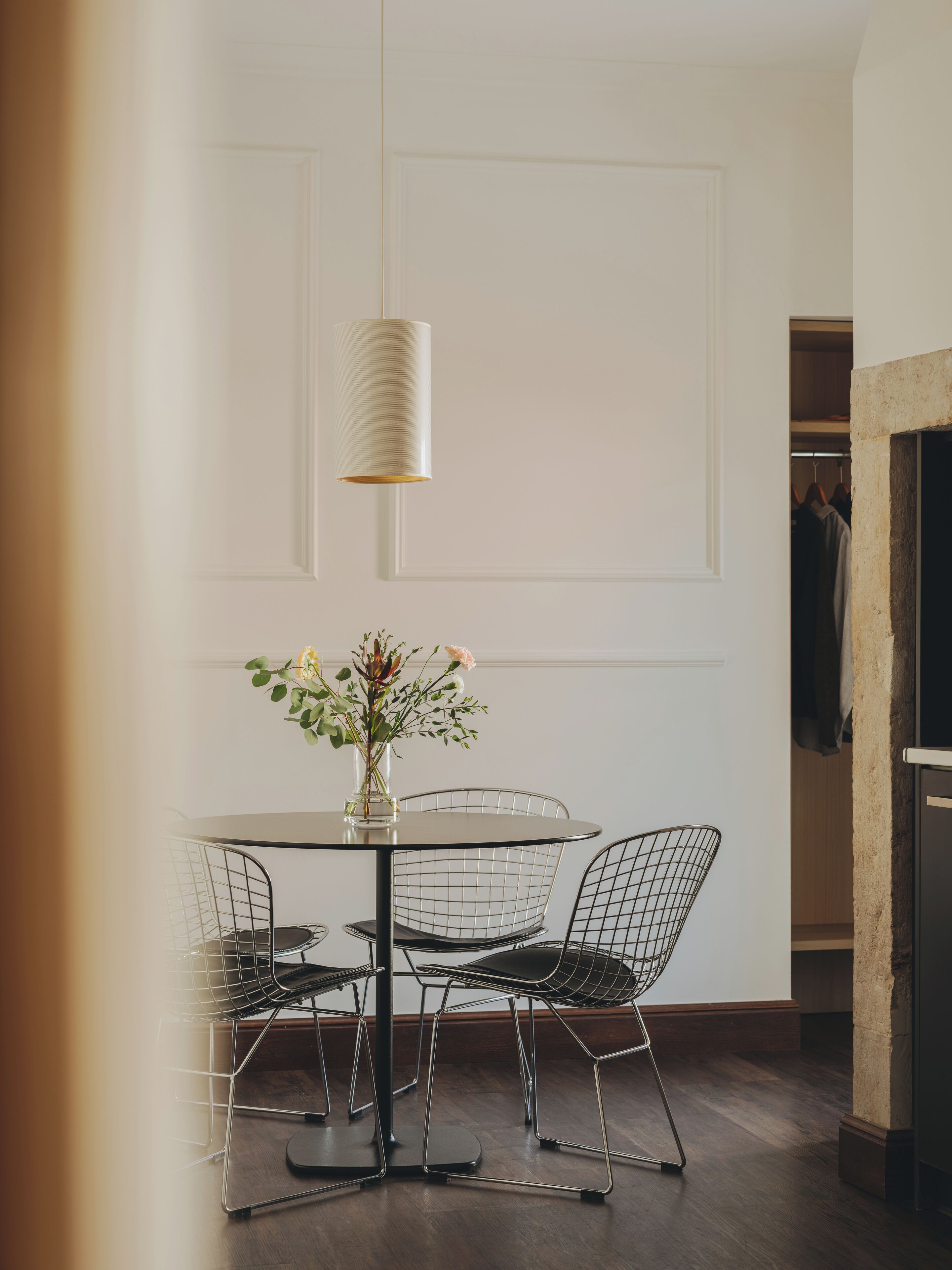 Et spiseområde med et rundt bord, stole med wirenet, en vase med blomster og en hængende lampe.