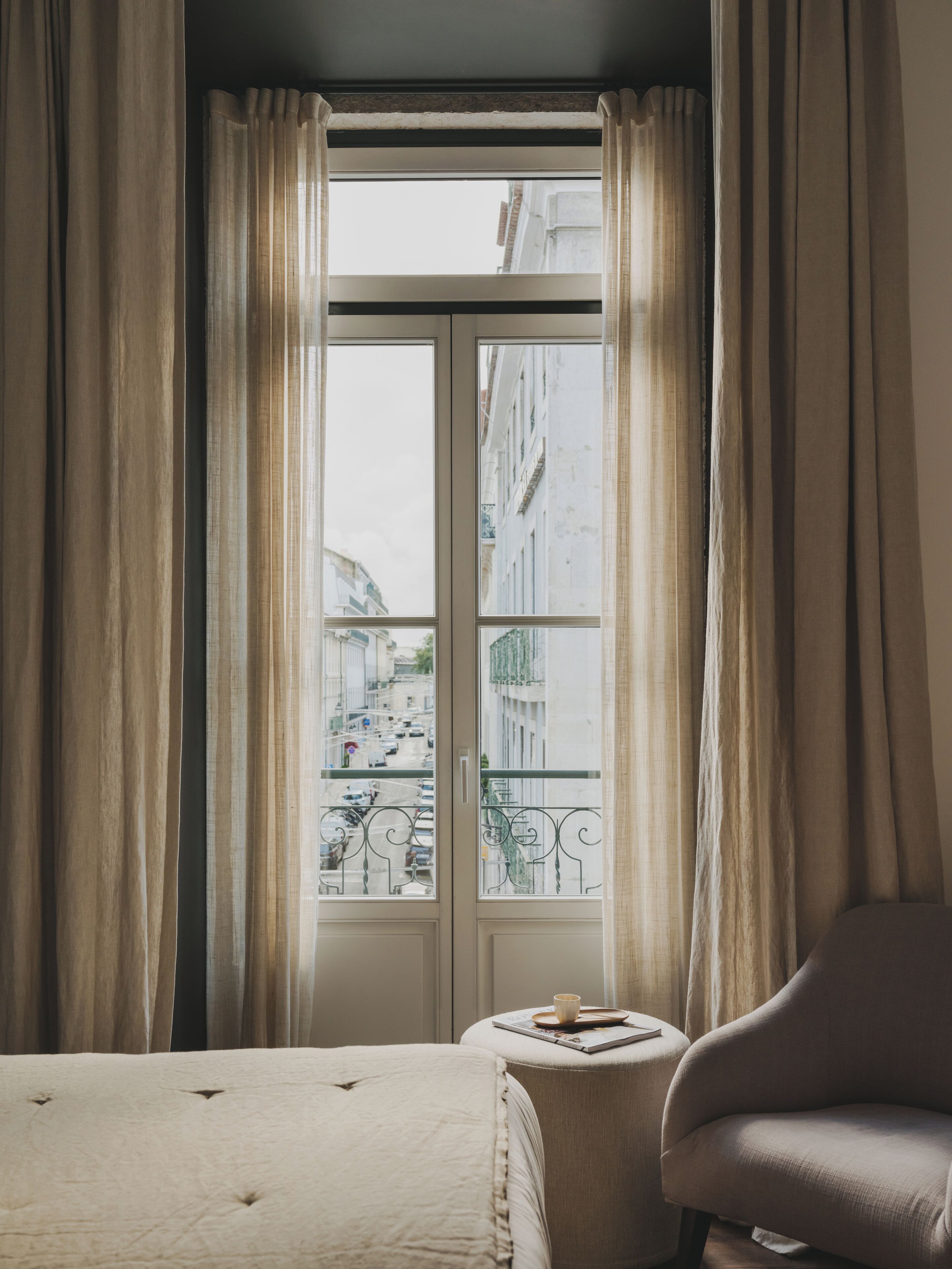 Una habitación de hotel con un ventanal grande, una cama, una silla y una mesita con una taza y un libro.